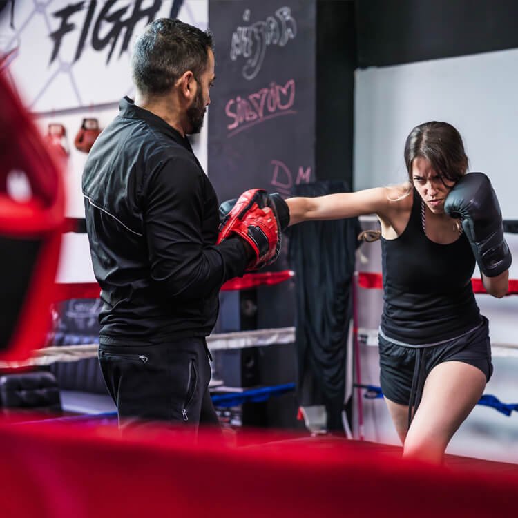 A female boxer in black tank top throws a punch at a coach wearing red boxing gloves inside a gym boxing ring.