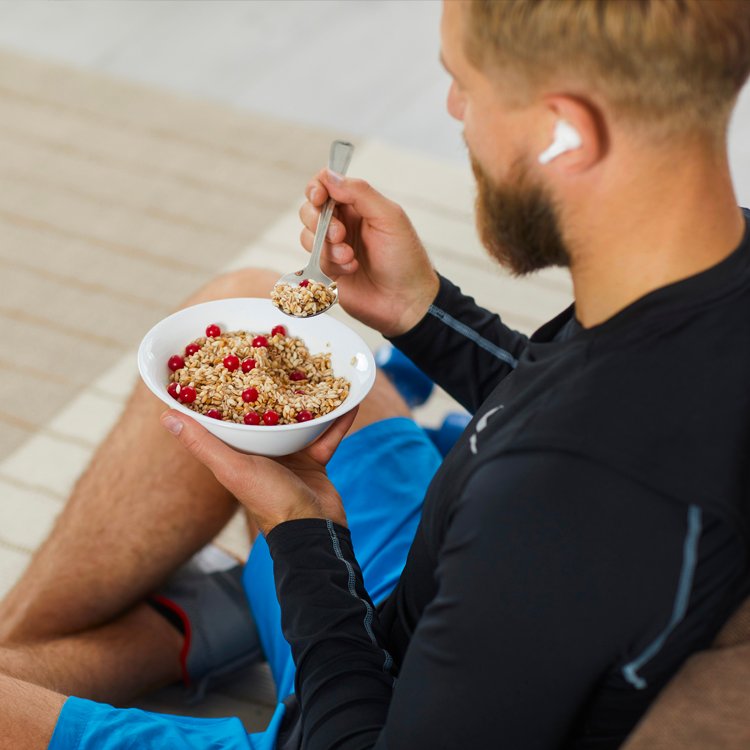 Man sitting outdoors, eating a bowl of granola with red berries, wearing a wireless earbud in one ear.