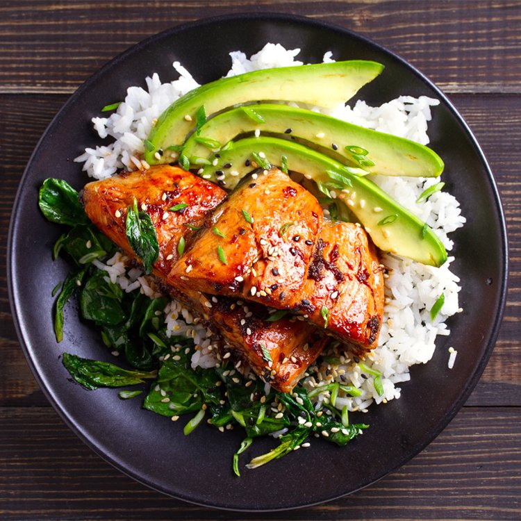 Glazed salmon fillets served over white rice with avocado slices, sautéed greens, and sesame seeds on a dark plate.