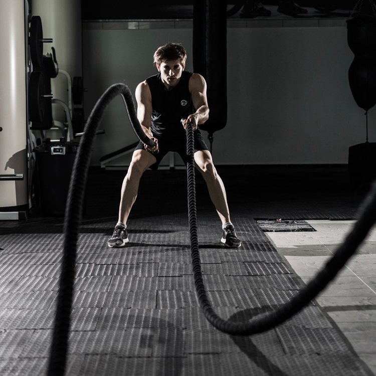 Man in a black tank top performs a battle rope exercise in a gym, knees bent, gripping the ropes as they whip forward.