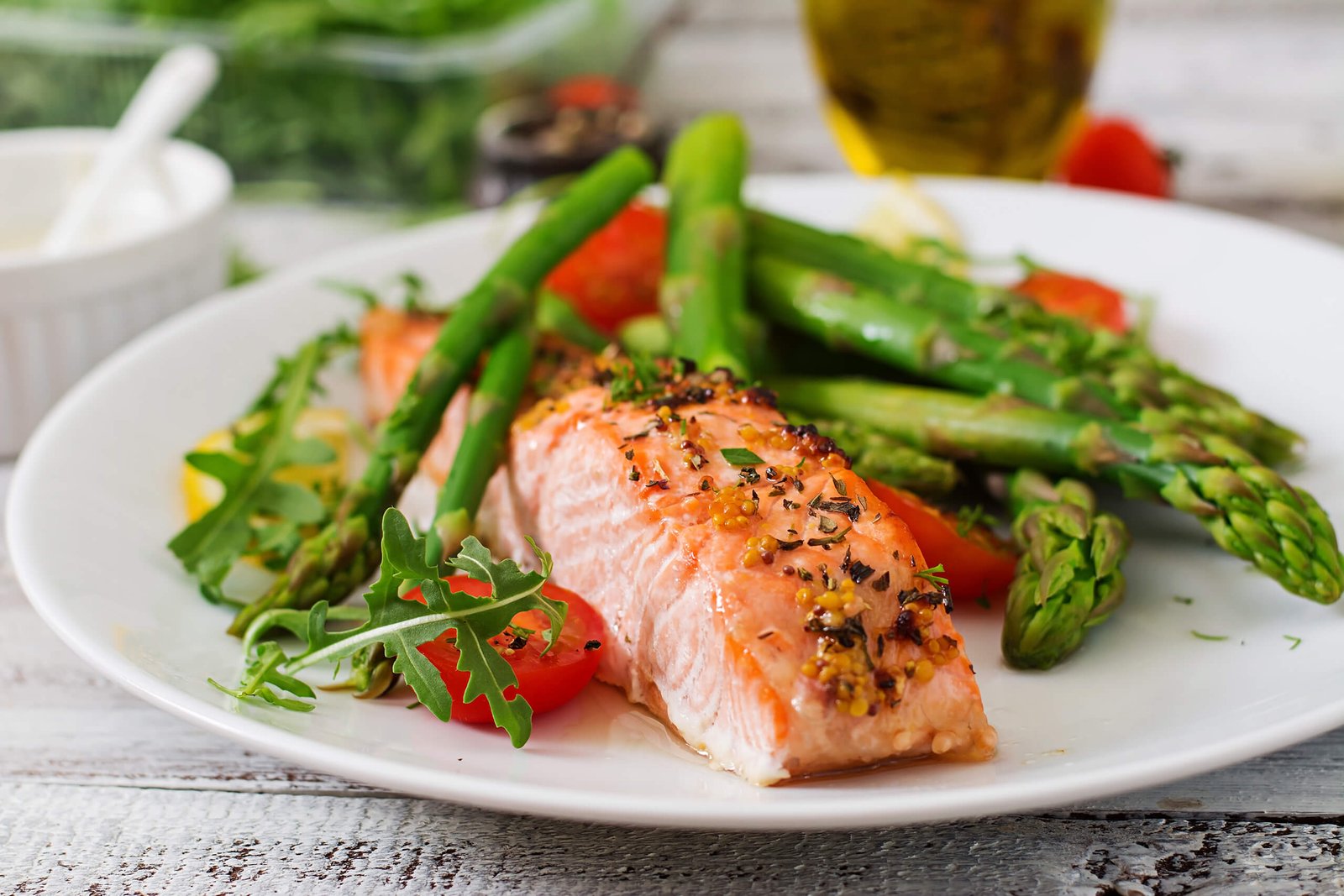 Salmon fillet with herbs on a bed of greens and asparagus on a white plate, with cherry tomatoes nearby.