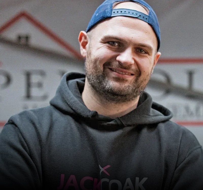Smiling man wearing a backward blue cap and dark hoodie with a logo on the chest, indoors.