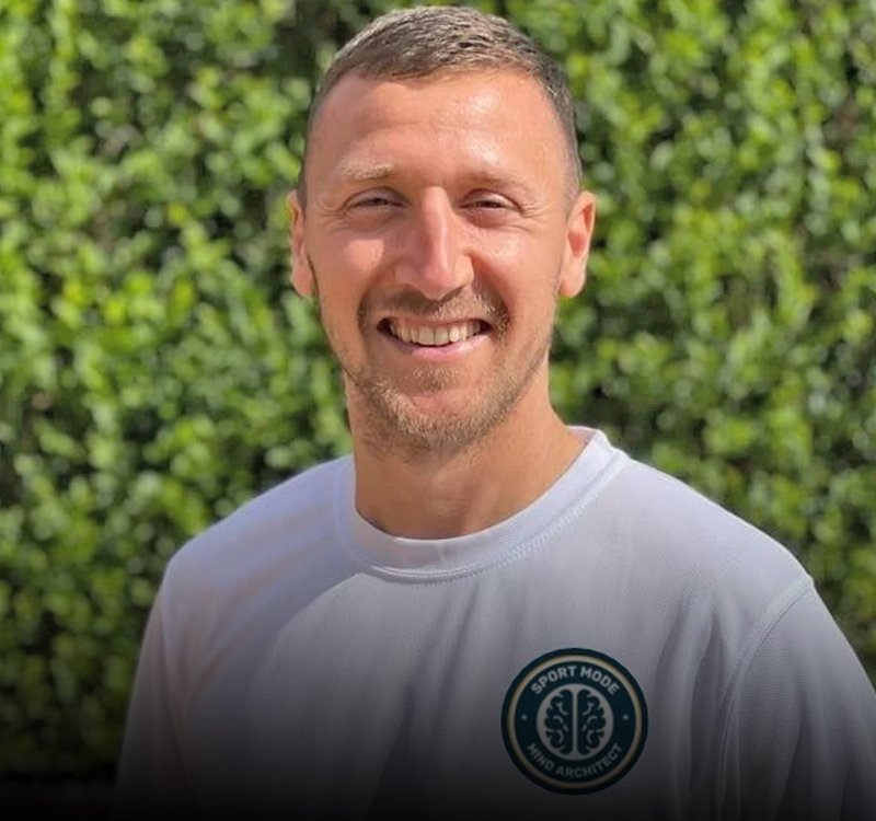 Portrait of a smiling man with short hair wearing a white T‑shirt, standing in front of a green hedge; circular logo visible on chest.