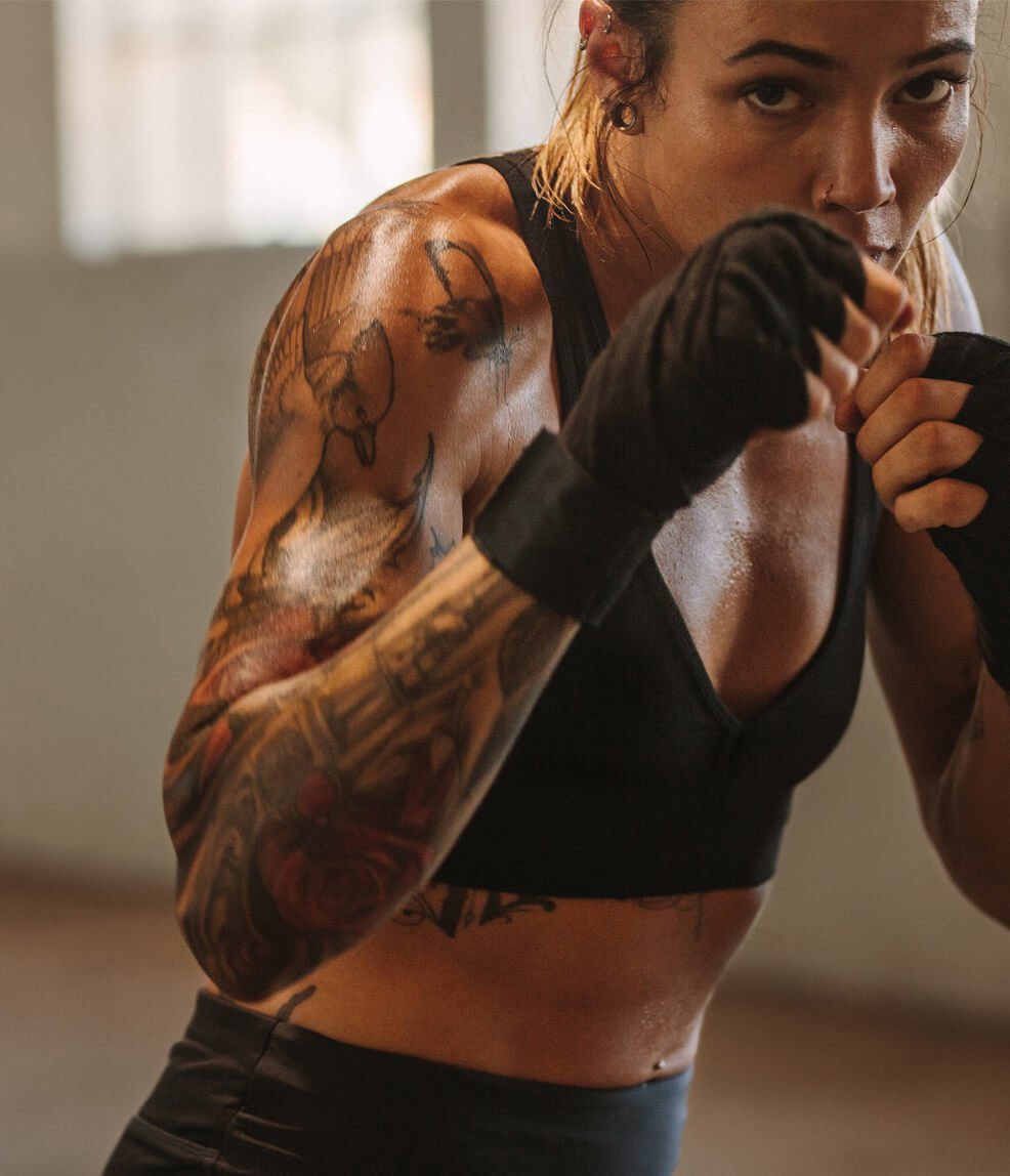 Female tattooed athlete wearing a black sports bra trains boxing, fists wrapped and raised in a focused stance.