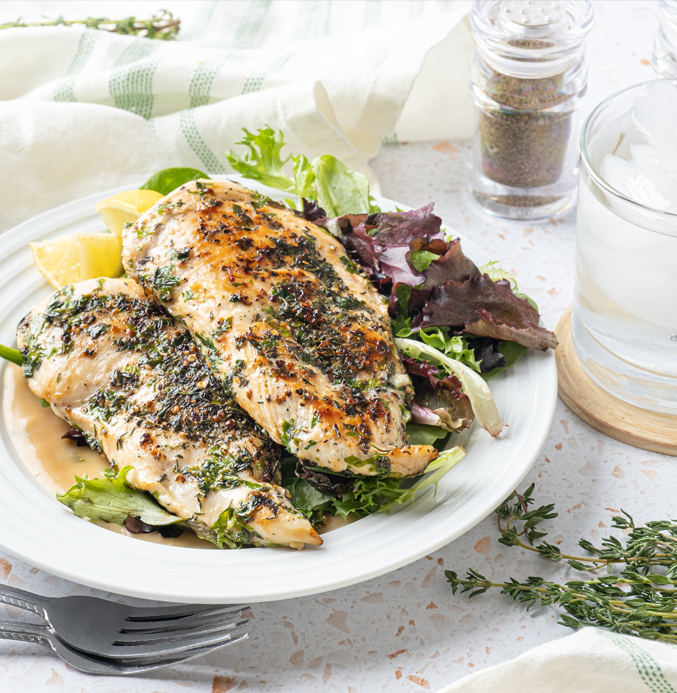 Grilled herb-marinated chicken breasts on a bed of mixed greens with lemon wedges on a white plate