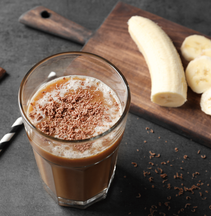 Glass of chocolate banana smoothie topped with chocolate shavings on a dark countertop, with a partially sliced banana on a wooden cutting board nearby.