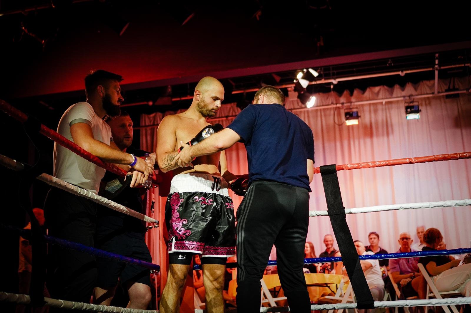 Boxer in floral trunks receives guidance from a trainer inside a boxing ring as spectators watch from the background.