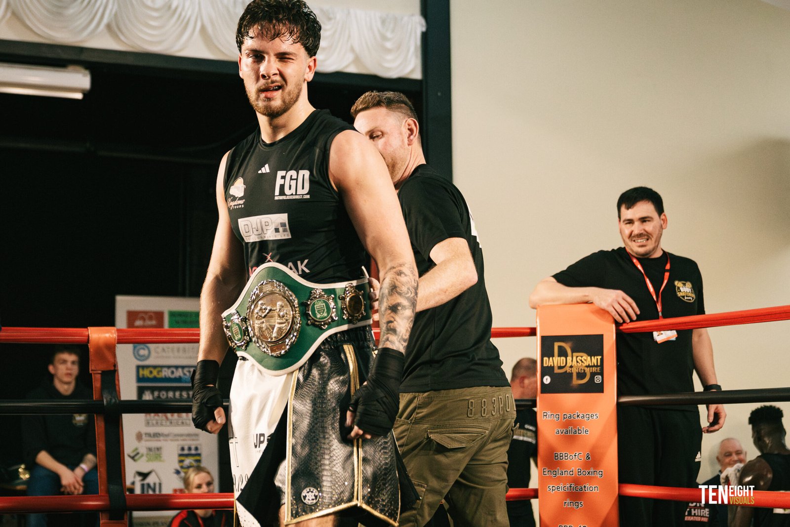Boxer wearing a large championship belt in a boxing ring, with team members nearby.