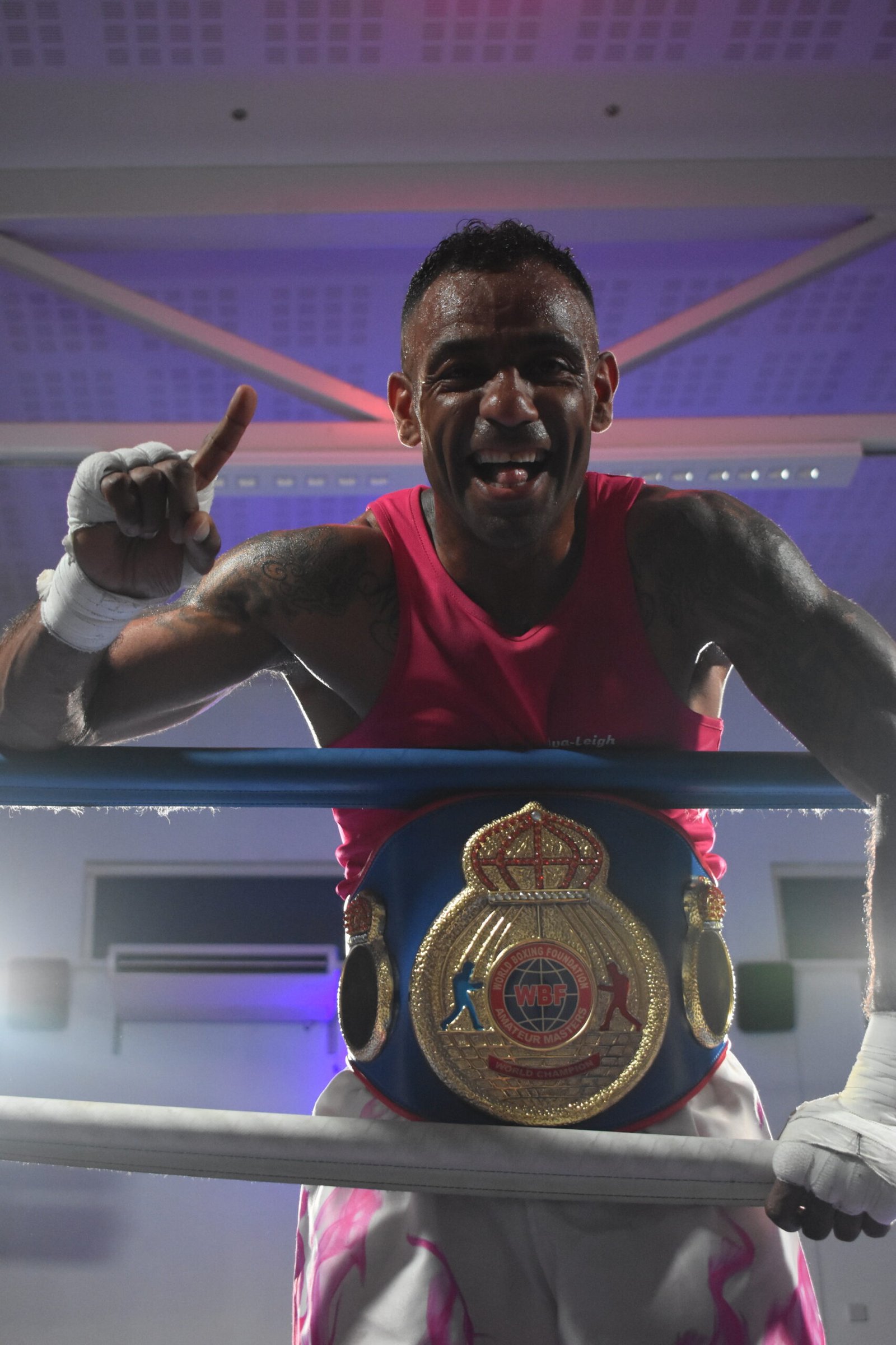 Smiling boxer in red trunks wearing a large championship belt in the ring, celebrating a win from the camera view.