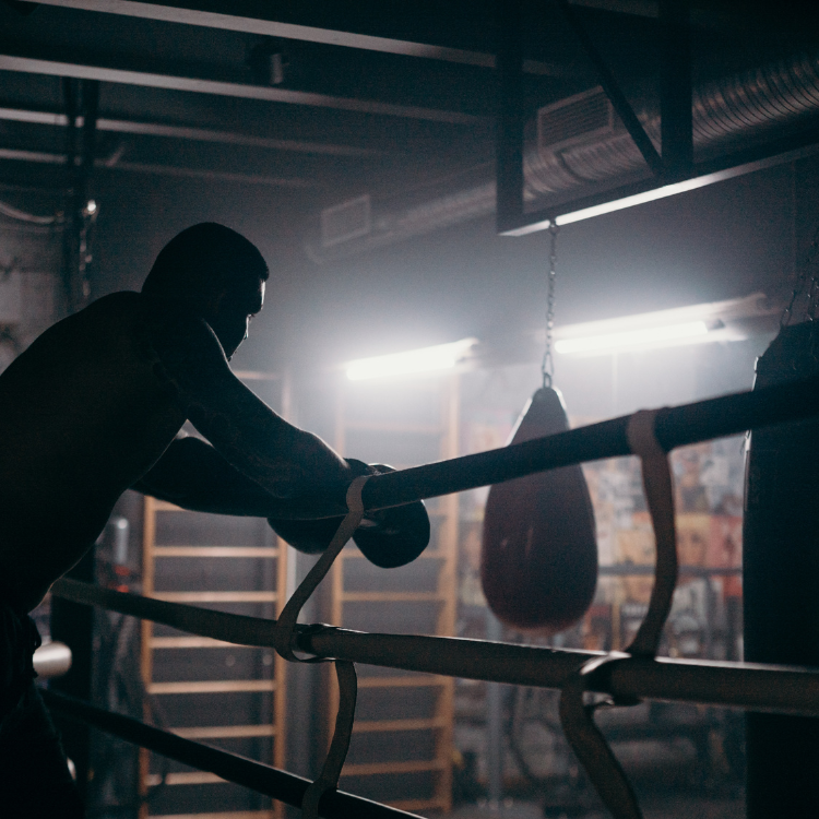 Boxer training in a dim gym, leaning on boxing ring ropes with a punching bag visible in the background.