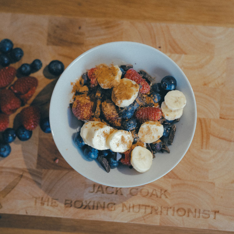 Bowl of granola with blueberries, raspberries, banana slices, and cinnamon on a wooden surface.