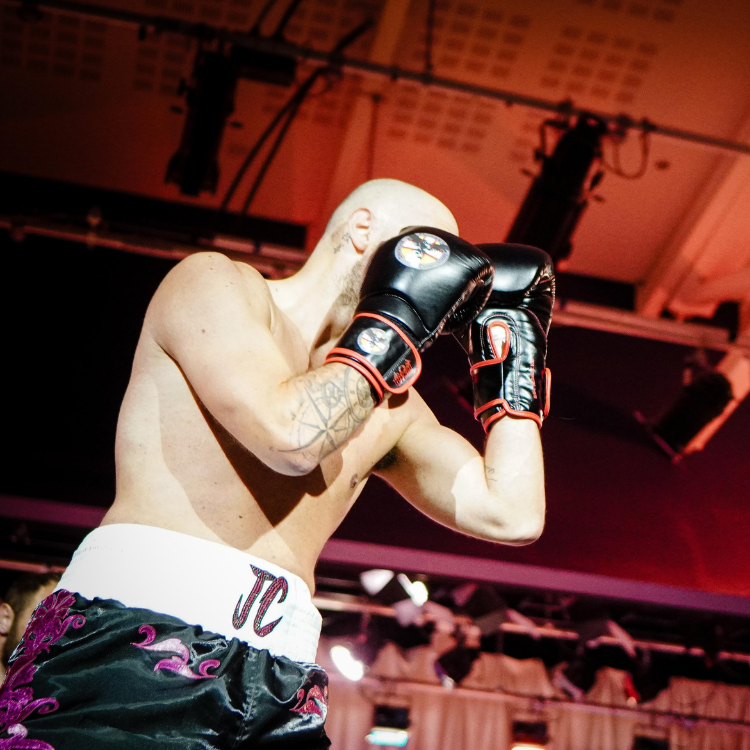 Male boxer wearing black gloves with red trim, mid-fight stance, shirtless, in a ring with purple-patterned shorts and a white waistband labeled 'JC'