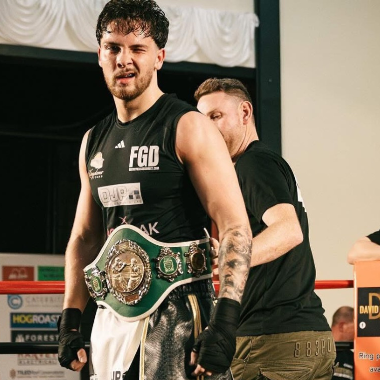 Boxer wearing a green title belt stands in a ring with a trainer behind him, after a win in a gym setting.