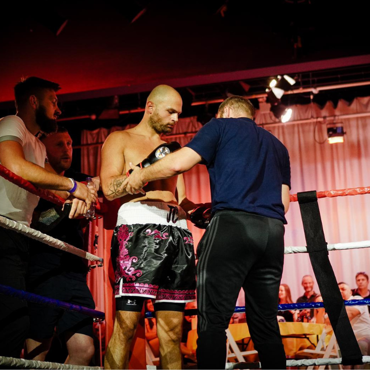 Boxer in colorful shorts stands in the ring as a trainer adjusts his stance and gloves.