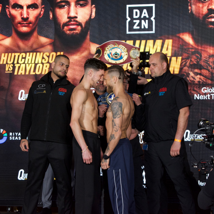 Two shirtless boxers face off inches apart as a championship belt is shown between them on a weigh-in stage with officials nearby.