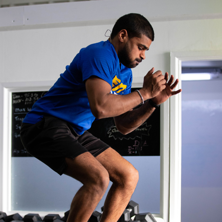 Man in a blue shirt performing a plyometric jump on gym boxes, knees bent and arms up for balance in a workout room.