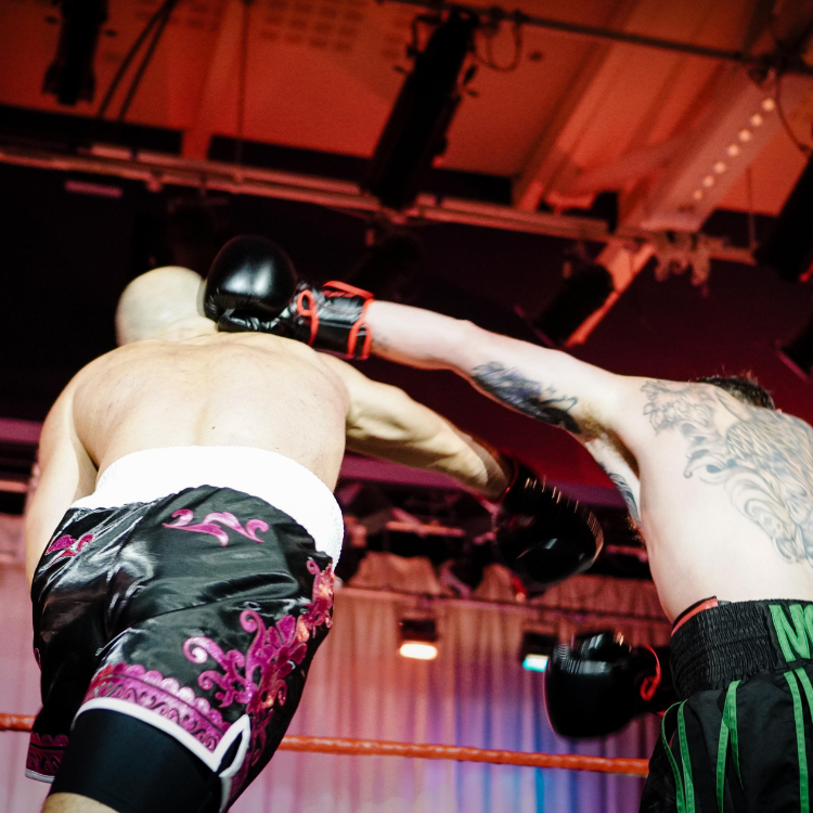 Two boxers clash in the ring as a punch lands; one wears black shorts with pink ornate patterns, the other has green-trimmed shorts and tattoos visible on his arm.