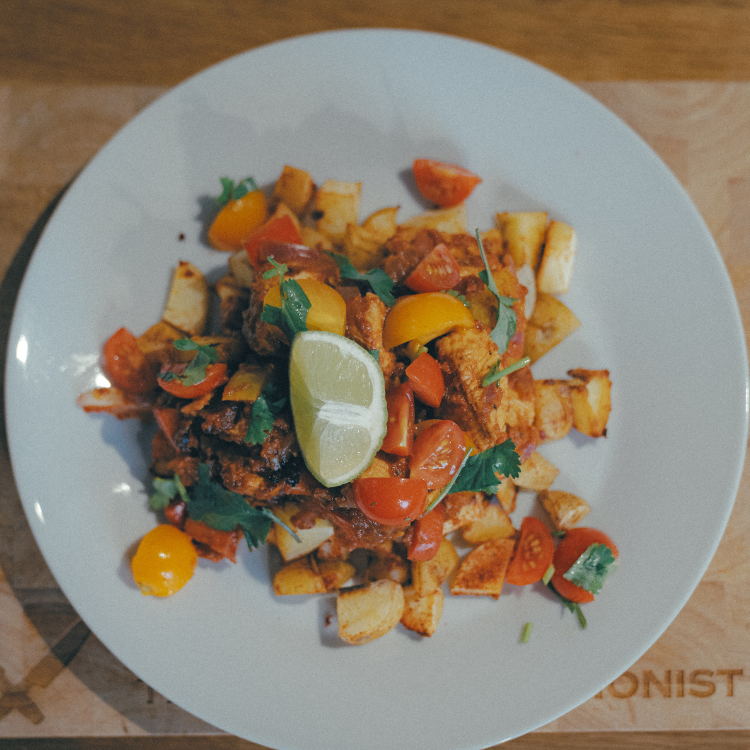 Plate of roasted potato cubes with chunks of meat, cherry tomatoes, cilantro, and a lime wedge on a white plate.