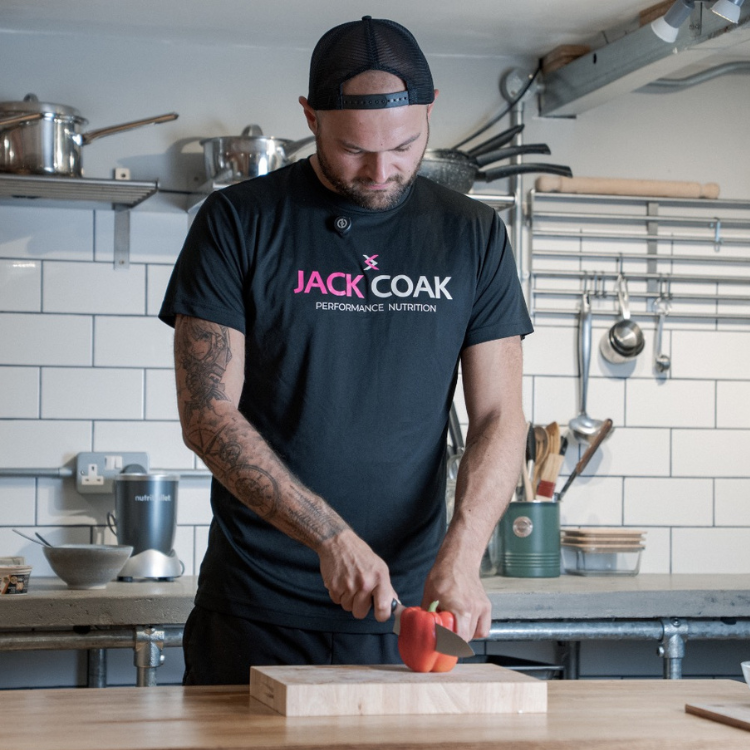 Man in a black JACK COAK T-shirt chops a red bell pepper on a wooden cutting board in a modern kitchen with tiled walls and metal shelves.