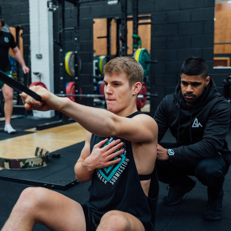 Young man sits on the gym floor performing a shoulder stretch with a resistance band, assisted by a trainer nearby.