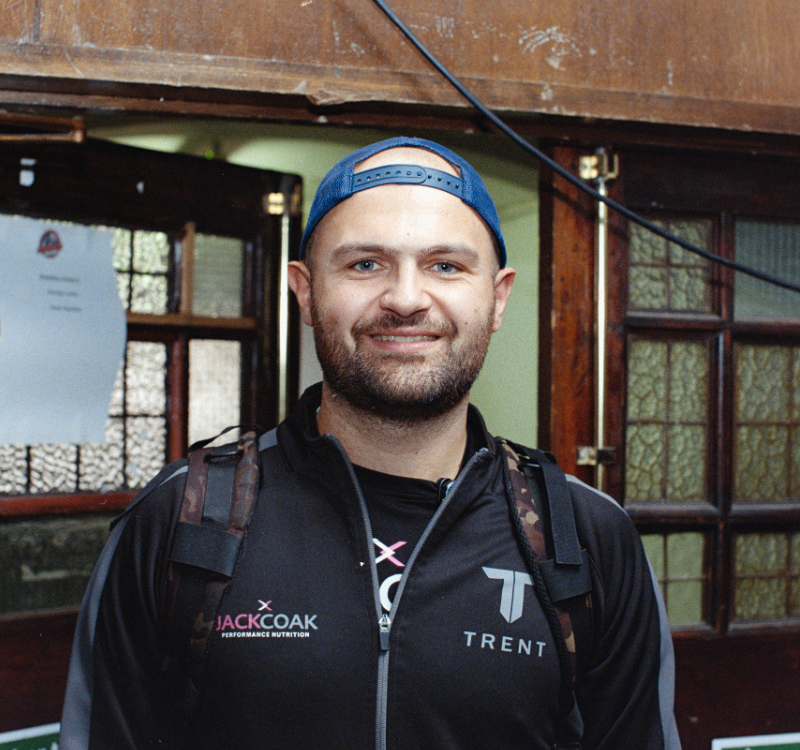 Smiling man wearing a blue backward cap and black jacket with Trent logo, indoors with wooden doors and a backpack strapped over the shoulders.